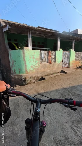 POV Video of Bicycle handlebars frame a quiet village street. Clear blue sky above a calm, sunlit road. Hand grips handlebars, suggest motion and purpose. Cables stretch overhead, connecting homes and