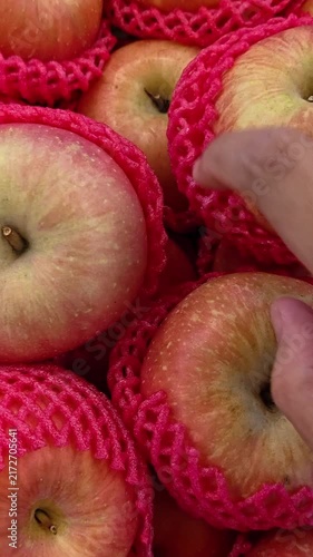 Vertical video of Hand selects a red apple from a pink netted bag. Apples glow with fresh, vibrant red skin. Basket holds more apples in soft mesh. Fruit displays warm, inviting market appeal. 4K POV 