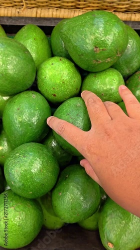POV Video of Hand selects ripe avocado from market basket. Firm, smooth green fruit gleams under soft light. Market scene suggests fresh produce, natural choice. Hand shows gentle care
