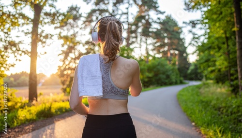 Young woman in sports bra and leggings walking on tree-lined path after outdoor run at sunset