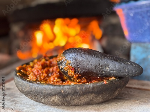 Traditional Indonesian stone mortar and pestle with freshly crushed red chili paste sambal. Rustic kitchen background with beautiful blurred fire bokeh. Authentic Asian culinary heritage concept.