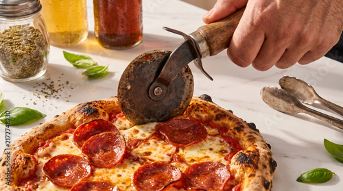 Obraz na płótnie Hand Slicing a Fresh Homemade Pepperoni Pizza with a Vintage Cutter on Marble Su