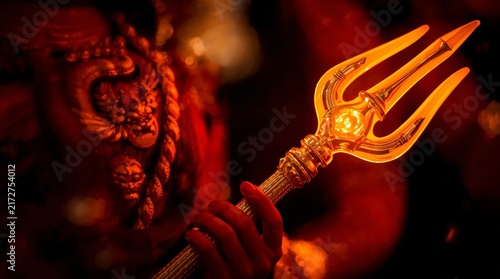 Close up of intricately carved deity idol gripping illuminated golden trident weapon with dramatic lighting against dark background showcases detailed close