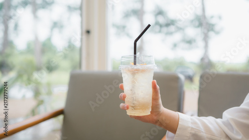 Person in white shirt holding a clear plastic cup of sparkling iced yuzu soda at a modern cafe. Conceptualizing relaxing weekend break, sweet summer beverage, cafe culture, and thirst quenching soda.