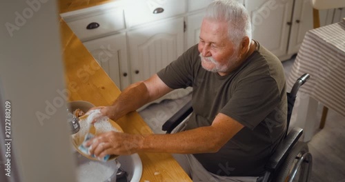 Independent senior man in a wheelchair washes dishes at the kitchen sink at home, managing his daily household routine on his own.