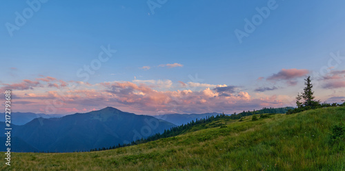 Canvastavla A vast panoramic view of grassy mountain slopes at sunset, featuring distant blue peaks and soft pink-tinged clouds floating in a clear sky