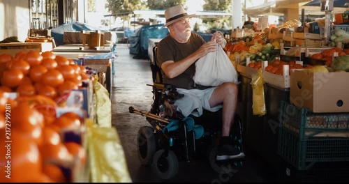 Female market vendor hands a reusable bag of fresh vegetables to a senior man in an electric wheelchair as he finishes shopping and says goodbye with a smile.