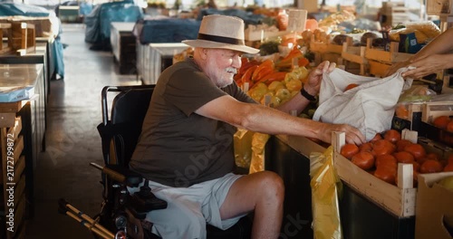 Senior man in an electric wheelchair selects fresh vegetables at a market stall and places them into a reusable bag while smiling and talking with a female vendor.