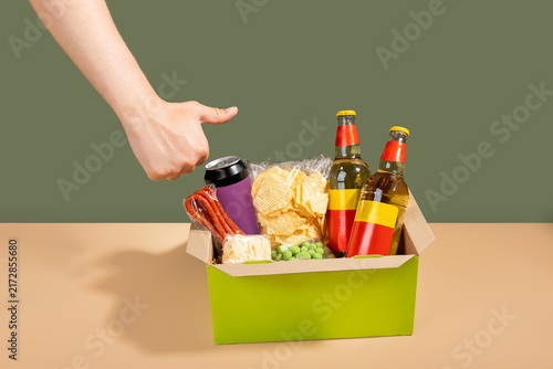 Hand pointing at a green subscription gift box filled with beer bottles, chips, and snacks. Monthly beer and snack delivery box presentation.