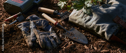 Gardening tool and glove beside tree watering bag on mulched soil, warm sunlight highlighting outdoor planting equipment and natural texture in garden setting