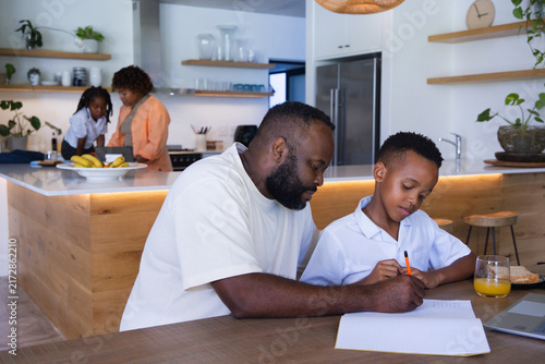 African American family working in home kitchen with father guiding youth son writing notebook