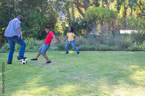 African father and two boys playing with soccer ball on backyard lawn near shrubs, copy space