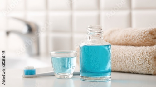 Blue mouthwash in clear glass bottle with small glass cup and toothbrush on white bathroom counter, folded beige towel, oral hygiene concept.