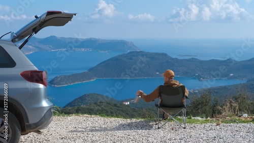 Tourist enjoying breathtaking sea. Traveler sitting on a camping chair, drinking from a thermos and admiring a beautiful seascape with islands and turquoise water, next to his car with open trunk