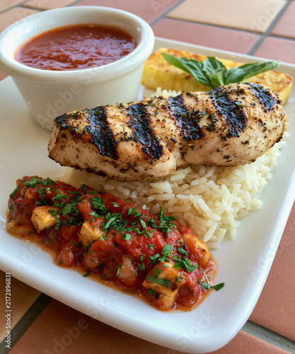 Grilled Chicken Breast with Rice and Sauce A succulent grilled chicken breast rests atop a bed of fluffy white rice, accompanied by a vibrant tomato salsa and a small bowl of rich marinara sauce.