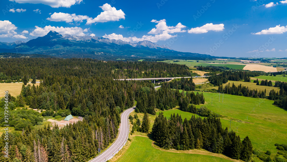 Fototapeta premium Mountains and green fields near a winding road in a rural area. A winding road runs through green fields and trees with mountains in the background.