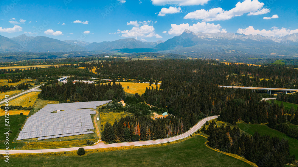 Naklejka premium Solar panels sit beside a highway in a rural area surrounded by mountains. Solar panels cover land near a highway, with mountains in the background and fields around.