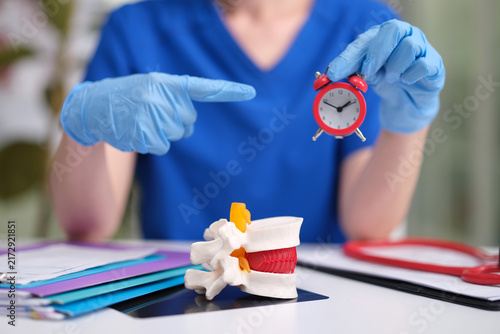 Photography A medical professional wearing blue scrubs and gloves points at a detailed spine model with an alarm clock in a clinic or office setting