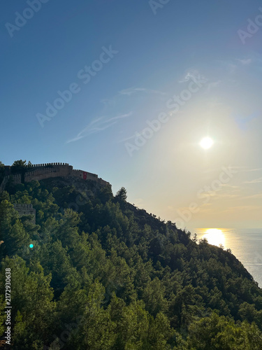 Scenic view of the historic Alanya Castle walls on top of a forested hill against a bright sun reflecting on the Mediterranean sea in Turkey
