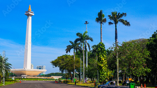 Fotografie The iconic National Monument (Monas) in Jakarta, Indonesia, is surrounded by a spacious plaza, green landscapes, palm trees, and modern city buildings beneath a bright blue sky
