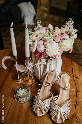 A bouquet of various and colorful flowers in a special package with two wedding rings lying on it, next to candles and high-heeled shoes on the wedding day