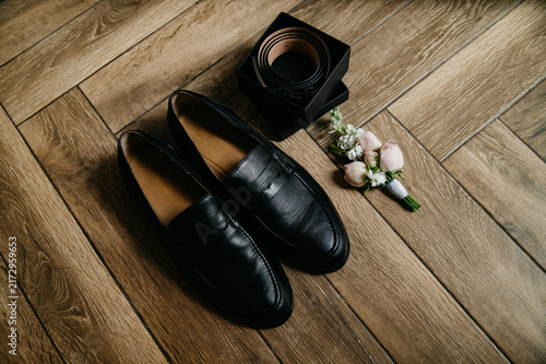A pair of classic black leather men's shoes next to a trouser belt and a boutonniere of flowers for the groom on his wedding day lie on a parquet background
