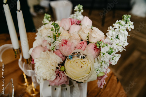 A bouquet of various and colorful flowers with two wedding rings lying on it on the wedding day for the bride and groom, and two candles standing beside it