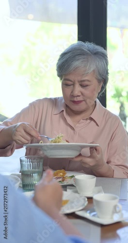 Senior asian retiree woman lifts pasta during reunion brunch at restaurant table, enjoying meal with close friend, relaxed dining, companionship, warm social bonding, Vertical lifestyle frame