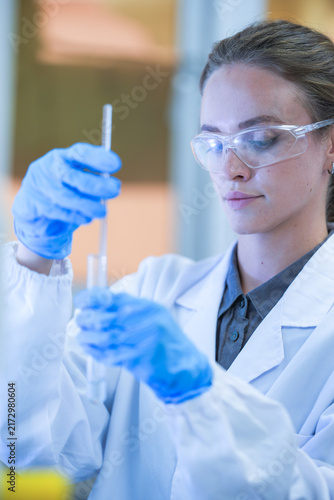 Fotografía A female research scientist in a white lab coat, hairnet and clear safety goggles looking straight at the tube contents at a well-equipped science laboratory bench
