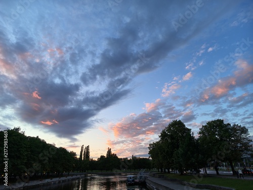 Wide dusk sky above tree lined park canal with dramatic blue clouds and soft pink sunset glow, calm reflections on water, dark green silhouettes, peaceful outdoor landscape