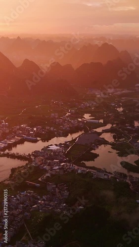Vertical Video: Golden sunlight breaking through rain over a riverside village with karst mountains in the distance, creating a misty glow