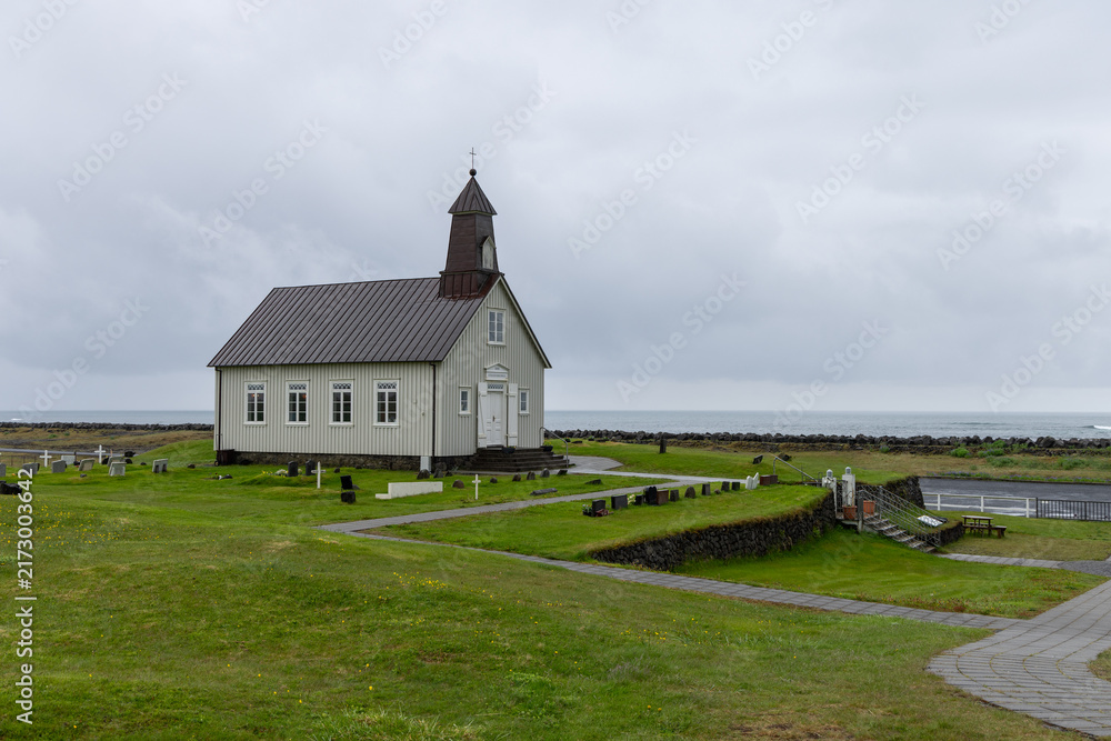 Fototapeta premium Strandarkirkja stands alone in low coastal grass at Selvogur, Iceland, beneath an expansive stormy sky. The white wooden church occupies the low horizon, emphasizing the scale of the open plain.