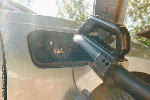 Closeup of electric vehicle charging plug inserted into car charging port, sun flare across frame, textured handle and cable visible, open hatch, metallic car body, clean outdoor background