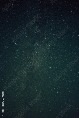 Long exposure vertical view of night sky showing bright Milky Way band, countless stars, soft nebula glow, deep teal-black tones, and faint meteor streak above quiet horizon, with night, sky, star