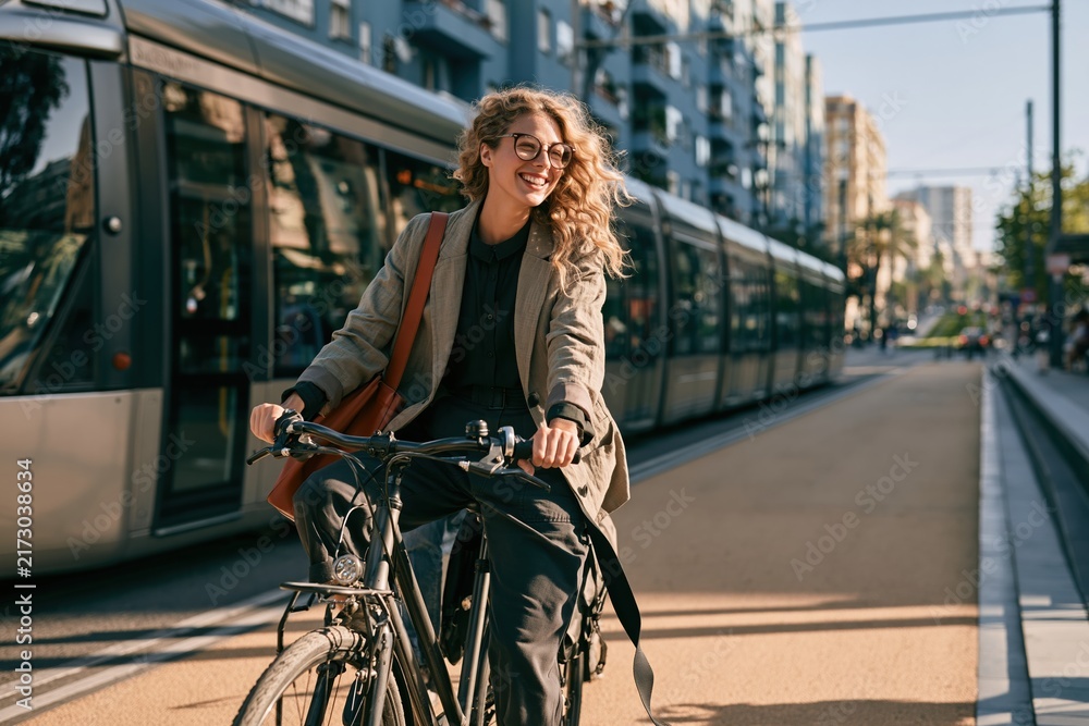 Obraz premium Young businesswoman riding a commuter bicycle on a dusty city street, smiling with a burnt orange bag for sustainable urban transport and eco-friendly travel