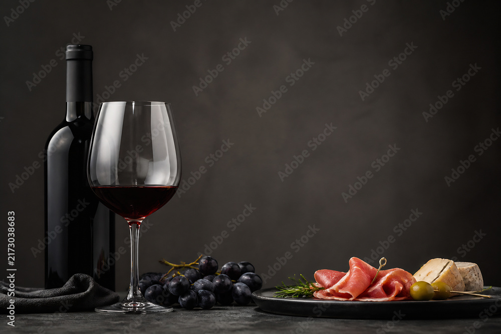 Fototapeta premium A bottle of red wine and a glass of red wine are displayed next to a platter of sliced cured meats, grapes, and cheese on a dark surface against a muted background.