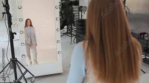 Beautiful young model with long hair wearing a stylish suit looking at her reflection in a large mirror with lights during a professional photoshoot in a modern studio backstage