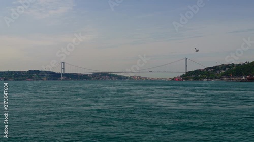 A wide landscape shot of the iconic suspension bridge crossing the Bosphorus strait in Istanbul with calm blue waters and distant hills under an overcast sky