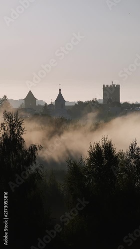 Vertical video. Mystical morning fog flowing through a valley with the silhouette of an ancient medieval town, castle, and church towers emerging from the mist against a warm sunrise sky and sunbeams