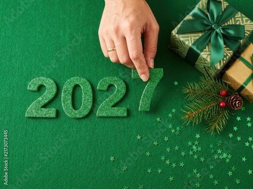 Hand placing a glittery green number 7 to complete the year 2027 on a green background with gifts and pine branch