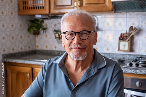 Senior gray-haired man with beard and glasses smiling warmly at the camera in a cozy domestic kitchen, head-and-shoulders portrait conveying contentment and healthy aging