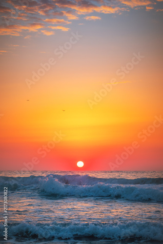 Obraz na płótnie Vertical shot of vibrant red sun rising over crashing ocean waves