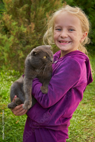 Smiling little girl holding cute scottish fold cat in hands outdoors, Cheerful kid girl playing with her pet cat on green grass background
