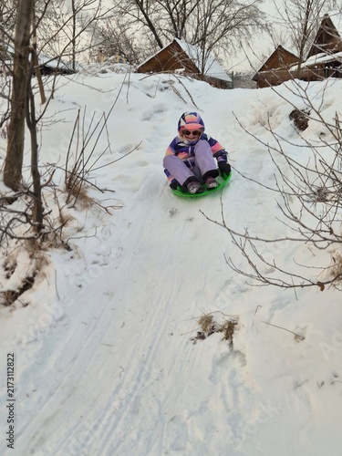 Canvastavla Cheerful girl sledding down a snowy hill on a green plastic saucer