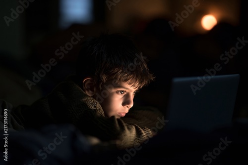 Young boy lying in bed at night looking at a glowing laptop screen in a dark bedroom setting