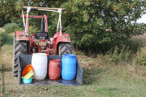 Red tractor transporting plastic barrels with water on a dry field. Concept image for water shortage, heatwave, and agricultural drought.