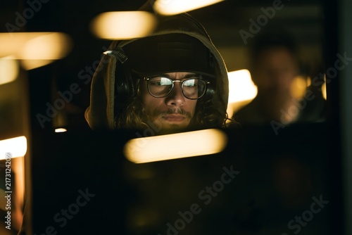 Young man wearing a hooded sweatshirt and glasses working intently at a computer in dark room