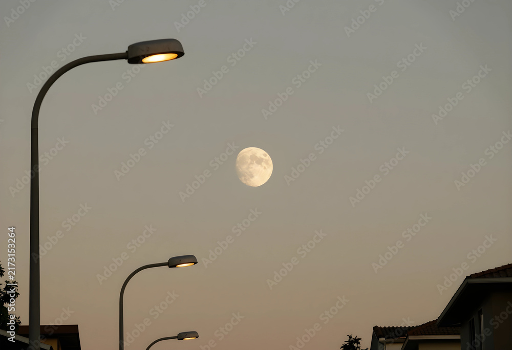 Obraz premium Evening atmosphere and urban twilight, full moon rising over suburban neighborhood with glowing street lights in a calm dusk sky for peaceful mood