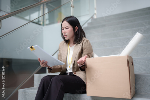 Bild auf Leinwand Disappointed businesswoman sitting on office stairs beside a cardboard box, looking frustrated and uncertain about career change, relocation, or workplace challenges