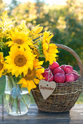 Beautiful rustic still life with apples in wicker basket and bouquet of sunflowers close up on table in garden, natural background. symbol of harvest, autumn season. September month calendar